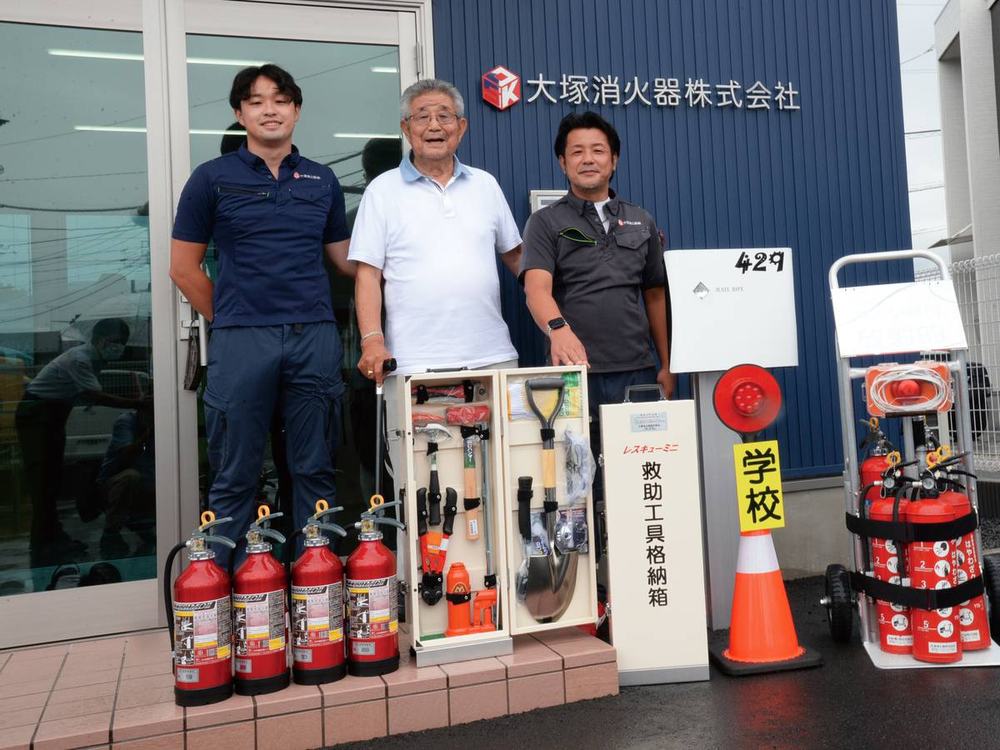 大塚消火器株式会社の社屋前に、レスキューシリーズの製品とともに並ぶ三代の経営陣